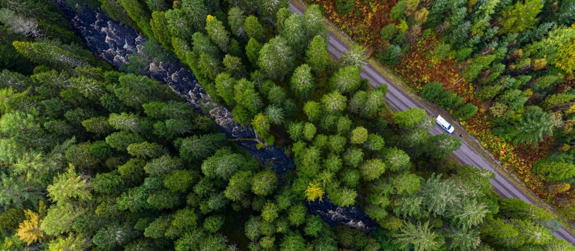 Vehicul văzut de sus care rulează pe o șosea paralelă cu un râu într-o zonă montană, împădurită