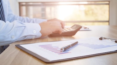 Mâinile unui angajat îmbrăcat în cămașă și cravată, care fac calcule cu un calculator portabil. Lângă calculator se află un clipboard cu grafice de business și un stilou așezat deasupra.