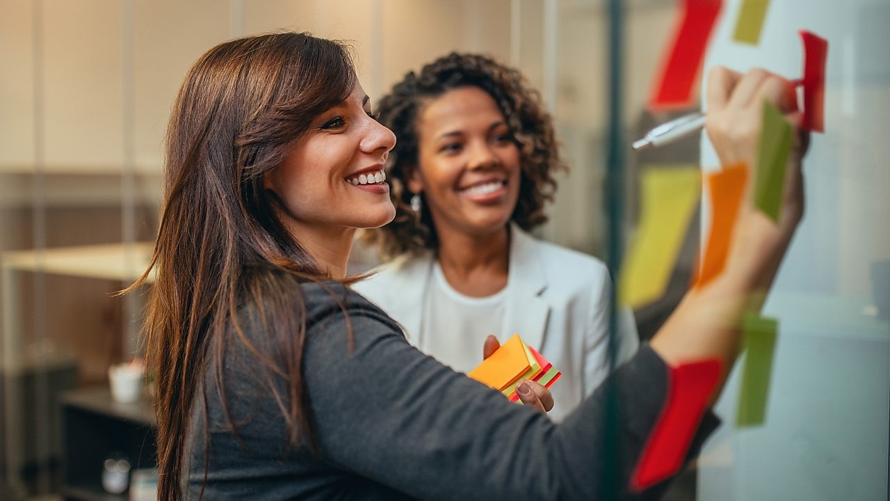 Două femei colege discută într-un birou modern. Una dintre femei scrie pe un post-it lipit pe un perete de sticlă, în timp ce cealaltă femeie zâmbește aprobator și citește.