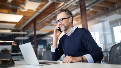 Mid adult business professional sitting at table with laptop and talking on mobile phone. Businessman working in office.