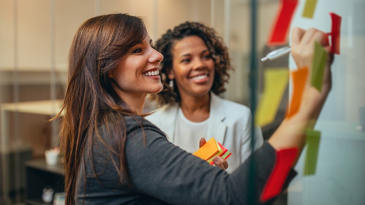 Două femei colege discută într-un birou modern. Una dintre femei scrie pe un post-it lipit pe un perete de sticlă, în timp ce cealaltă femeie zâmbește aprobator și citește.