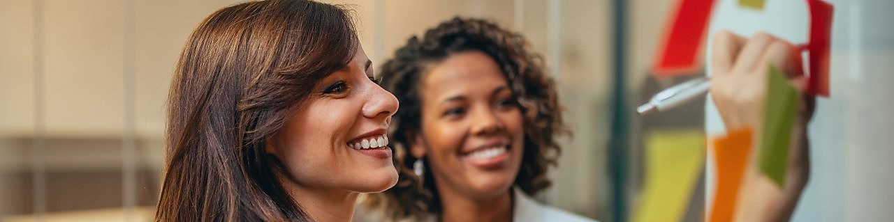 Două femei colege discută într-un birou modern. Una dintre femei scrie pe un post-it lipit pe un perete de sticlă, în timp ce cealaltă femeie zâmbește aprobator și citește.