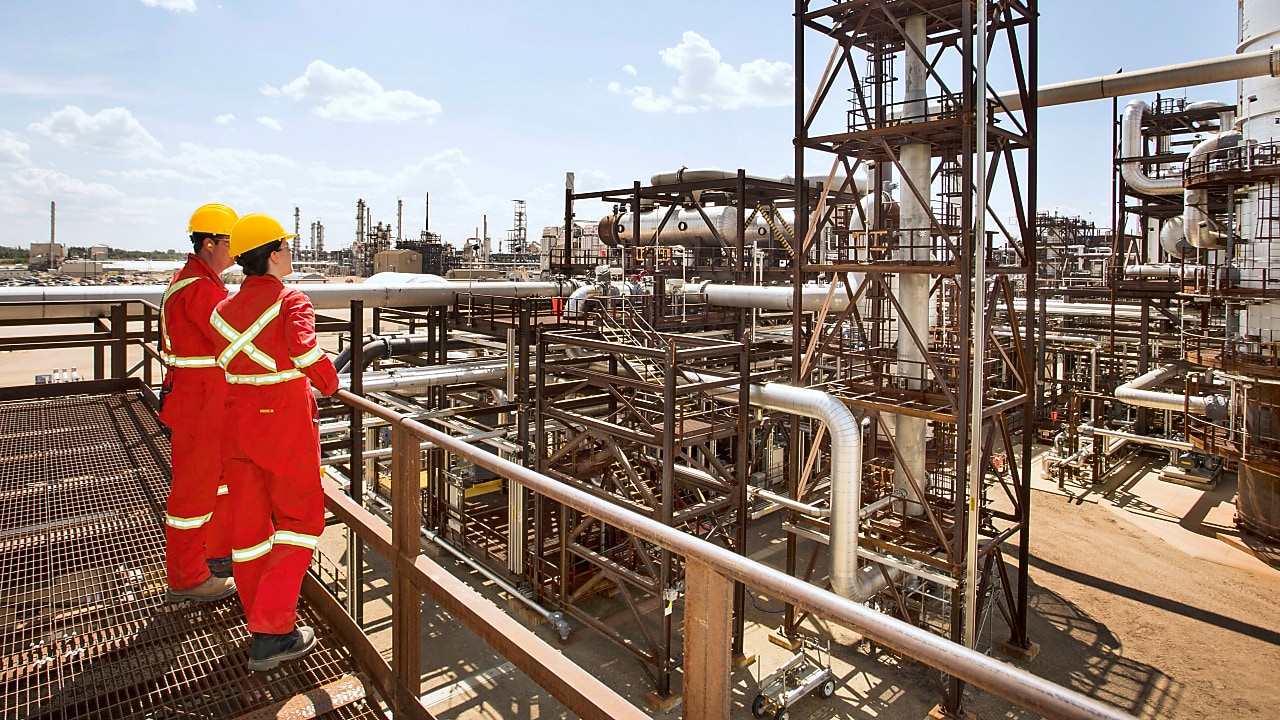 Muncitori cu combinezoane roșii cu benzi reflectorizante și căști de protecție galbene care discută pe schela unei rafinării