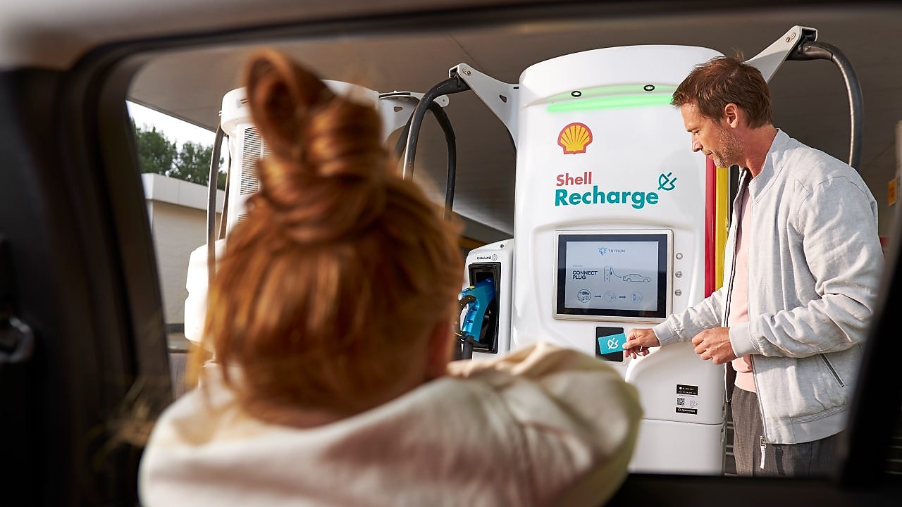 Un bărbat utilizând o stație de reîncărcare Shell în timp ce o fată se uită din mașină.