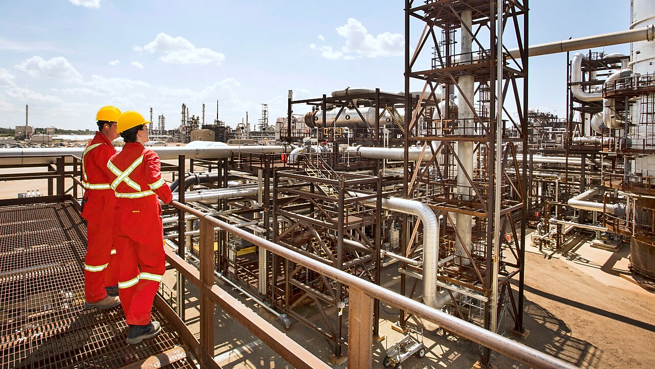 Muncitori cu combinezoane roșii cu benzi reflectorizante și căști de protecție galbene care discută pe schela unei rafinării