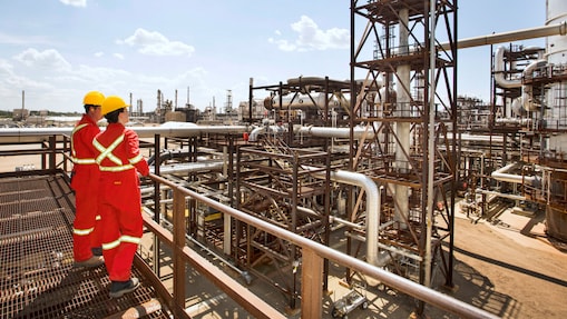 Muncitori cu combinezoane roșii cu benzi reflectorizante și căști de protecție galbene care discută pe schela unei rafinării