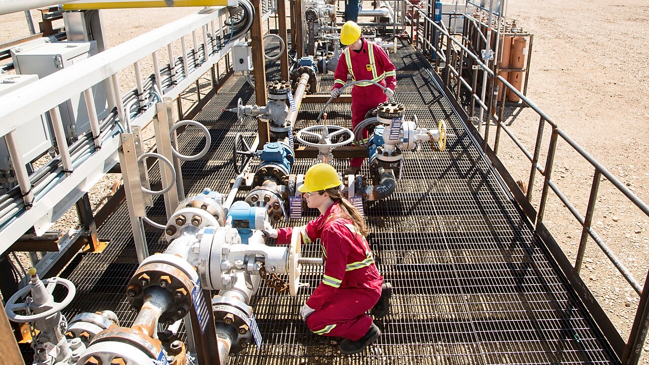 Femeie muncitor cu combinezon roșu și cască de protecție galbenă care controlează presiunea din conductele unei rafinării. Un al doilea muncitor îmbrăcat la fel care lucrează în plan secund.