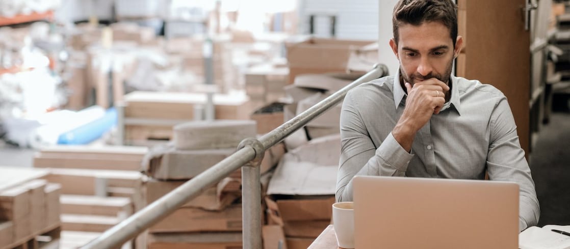 Angajat al unui depozit logistic care studiază ecranul unui laptop și își ia notițe într-o agendă. În fundal, cutii și ambalaje de mari dimensiuni așezate suprapus.