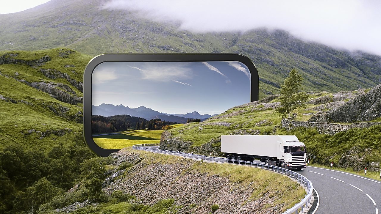 Camion alb care urcă o șosea unduitoare de munte, pe o vreme înnorată.