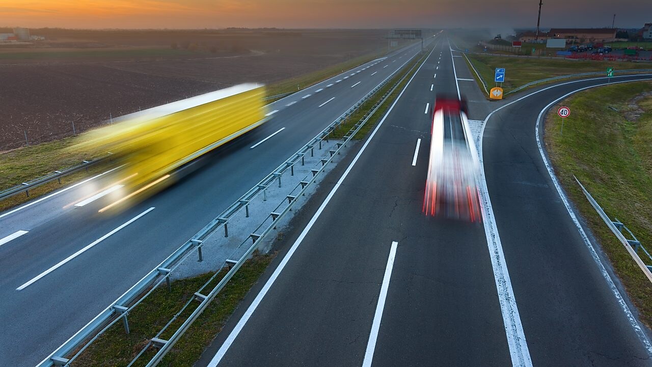 Două tiruri se deplasează pe o autostradă pe timpul unui frumos apus de soare; imaginea lor este ștearsă din cauza mișcării