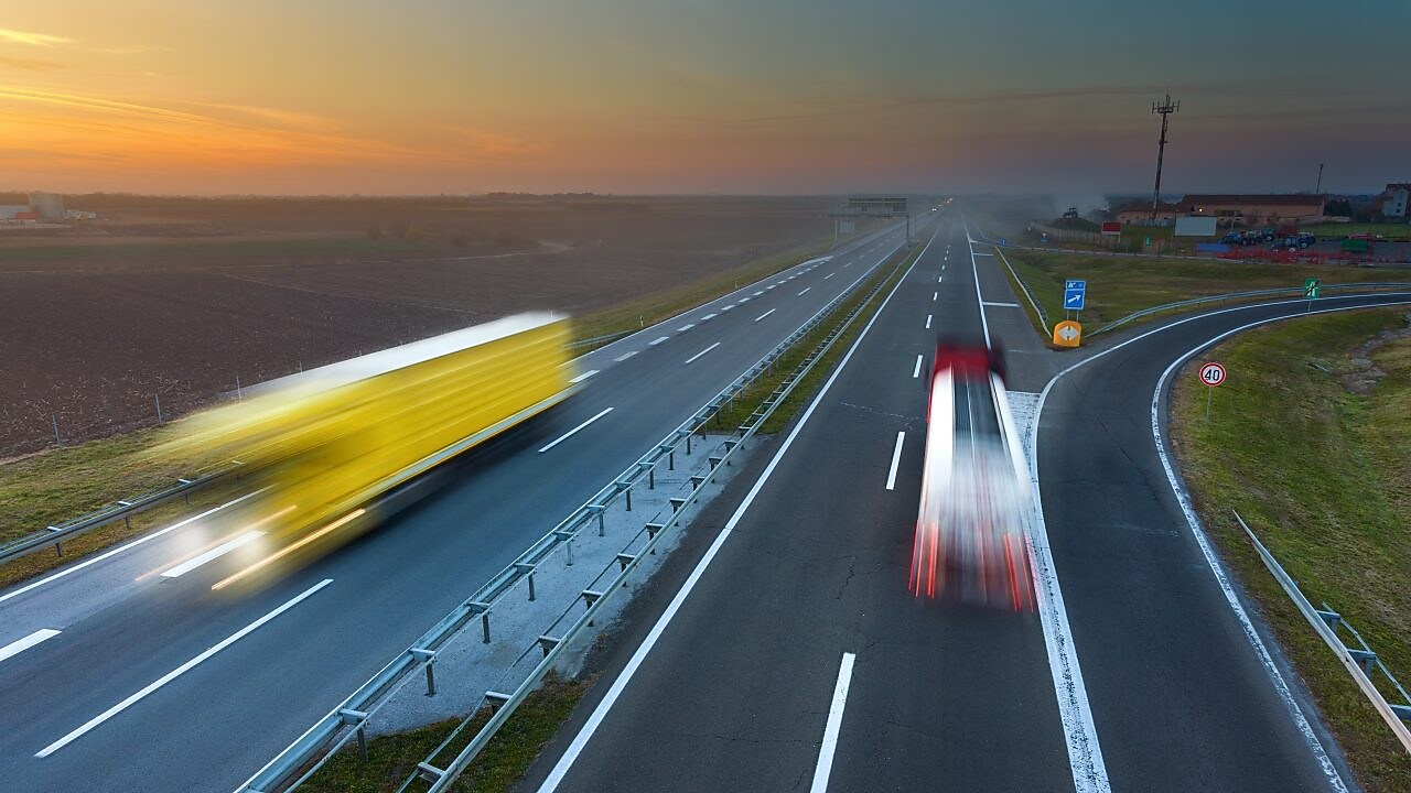 Două tiruri se deplasează pe o autostradă pe timpul unui frumos apus de soare; imaginea lor este ștearsă din cauza mișcării