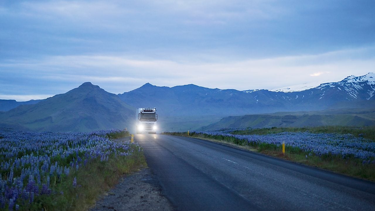 Camion cu farurile aprinse care se apropie pe o șosea de altitudine din golul alpin la orele dimineții. De o parte și de cealaltă a șoselei cresc flori alpine, iar în plan îndepărtat sunt creste montane înzăpezite.