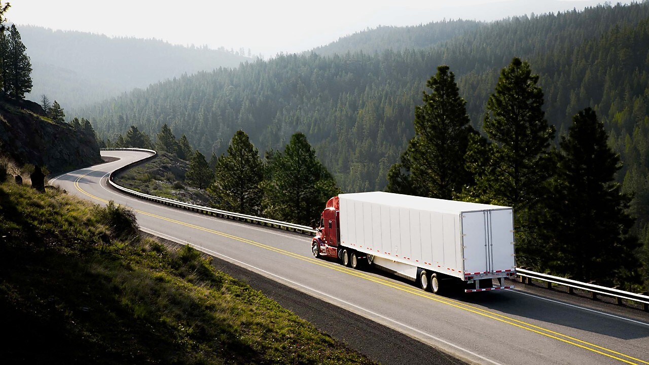 Camion care rulează pe un drum șerpuitor dintr-o zonă de munte, cu păduri de conifere.