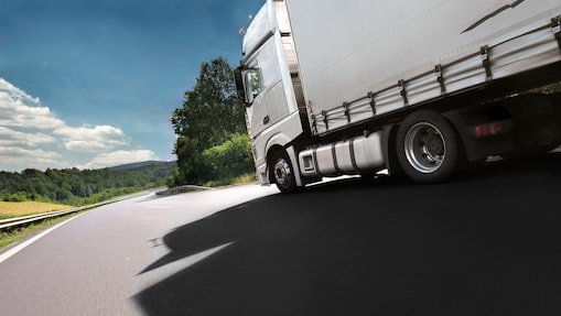 Vedere laterală a părții din față a unui camion alb care rulează pe o șosea dintr-o zonă de deal, cu păduri de foioase.