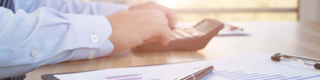 Mâinile unui angajat îmbrăcat în cămașă și cravată, care fac calcule cu un calculator portabil. Lângă calculator se află un clipboard cu grafice de business și un stilou așezat deasupra.