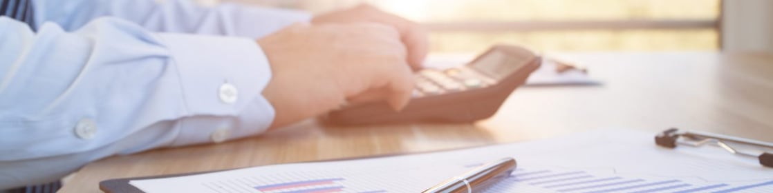Mâinile unui angajat îmbrăcat în cămașă și cravată, care fac calcule cu un calculator portabil. Lângă calculator se află un clipboard cu grafice de business și un stilou așezat deasupra.