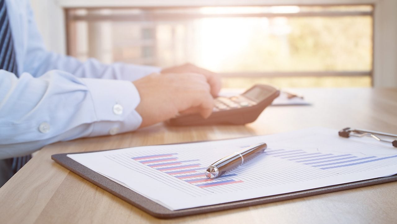 Mâinile unui angajat îmbrăcat în cămașă și cravată, care fac calcule cu un calculator portabil. Lângă calculator se află un clipboard cu grafice de business și un stilou așezat deasupra.